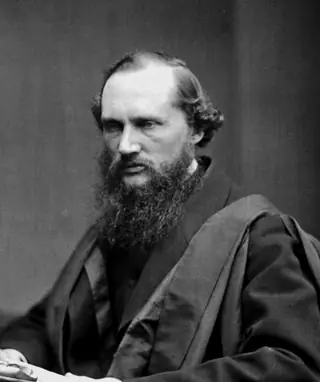 William Thomson in his forties, with a full dark beard, seated in an academic gown and holding papers.