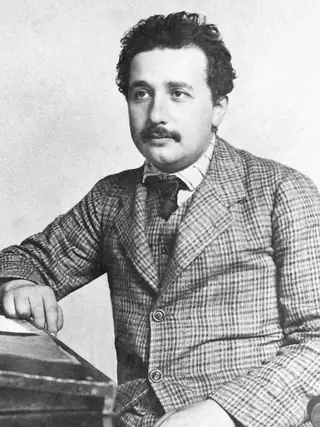 Albert Einstein in his mid-twenties, in a checked suit, seated with one arm on a sloping desk.